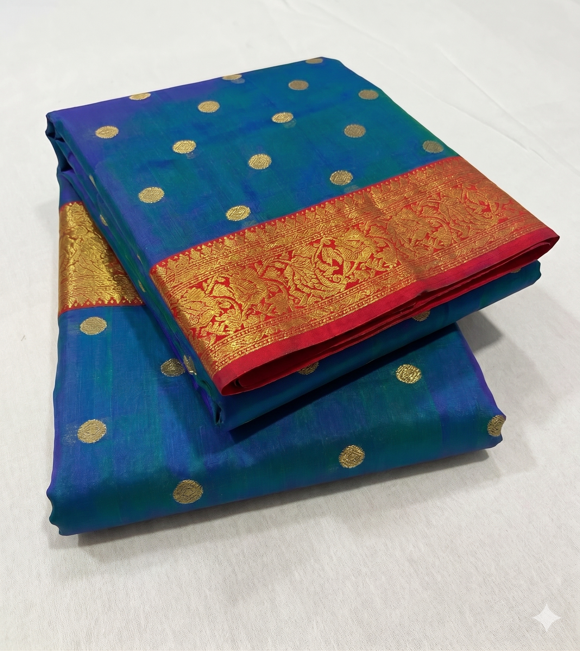 Neatly folded Royal Peacock Blue Chanderi Katan Silk Saree, providing an overview of the all-over gold coin butti pattern and the striking crimson red zari border. Authentically handloom
